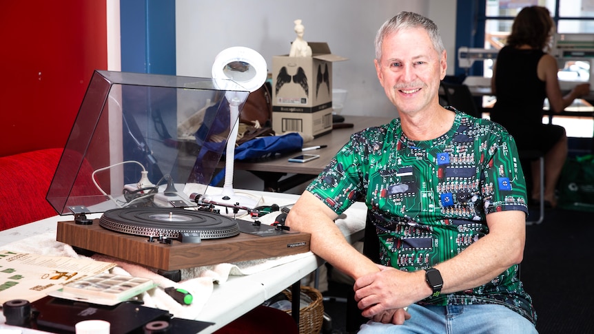 A man in a shirt with circuits printed on it smiles as he repairs a record player.