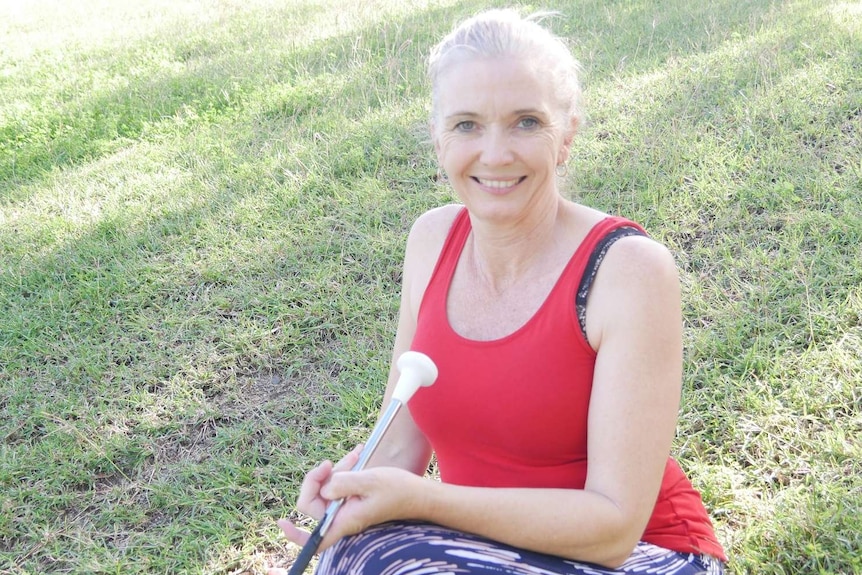 A woman with blonde hair in a red shirt sits on the grass and smiles at the camera.