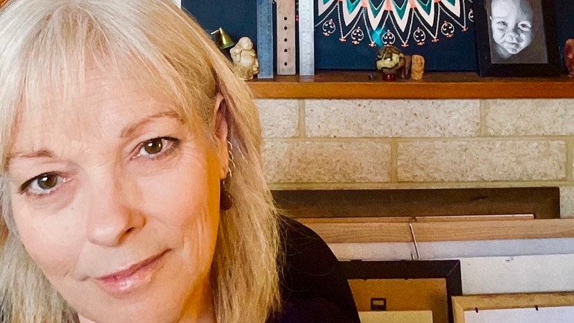 An attractive blonde woman in her early 60's stands in front of a mandala art work hanging on the wall behind her