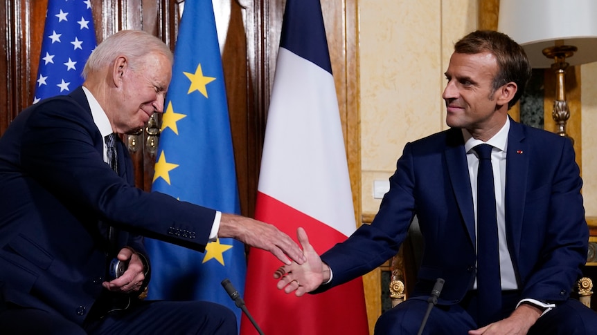 Mr Biden and Mr Macron shaking hands while seated in front of US and French flags