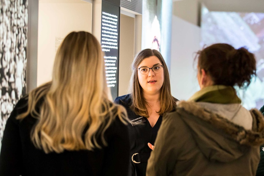 A woman faces the camera, explaining an exhibition in a museum to a group of people with their backs to the camera.