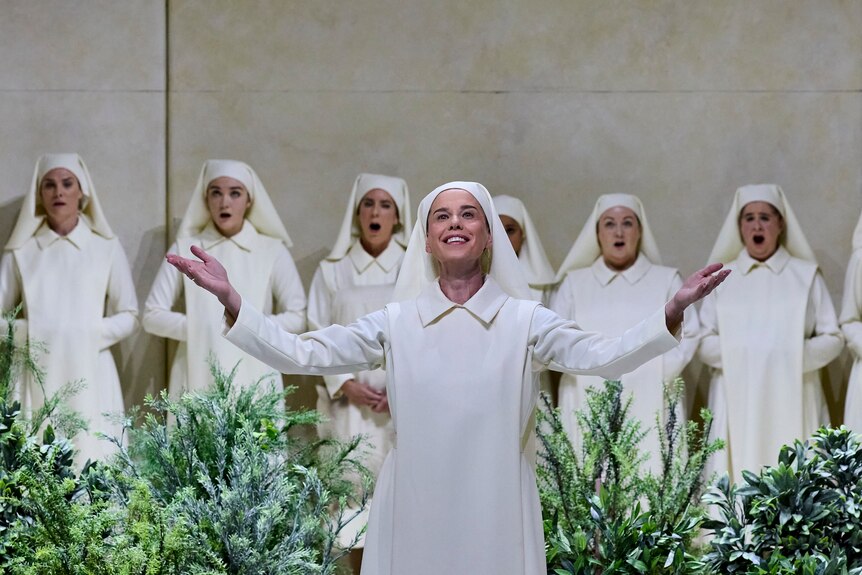 A nun dressed in all white stands arms open in front of plants and more nuns
