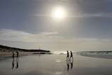 People walk across the wet sand at a beach as a strong morning sun beats down above them.