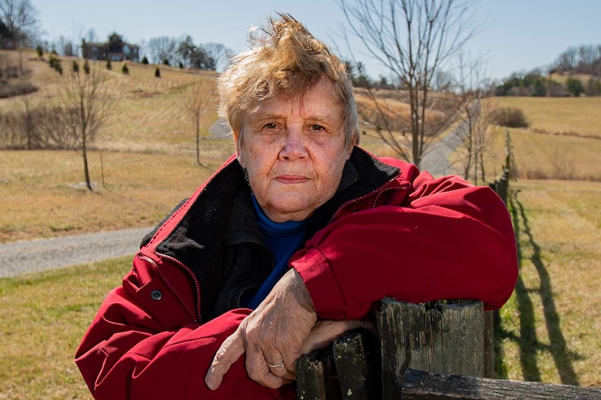 A woman in a red coat looking grim, leaning on a fence post