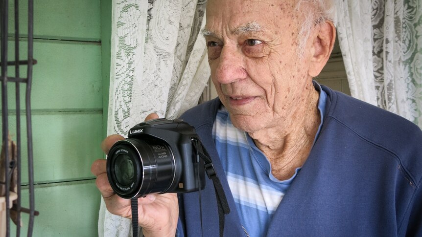 An elderly man holds a camera in one hand, a window with lace curtains behind him.