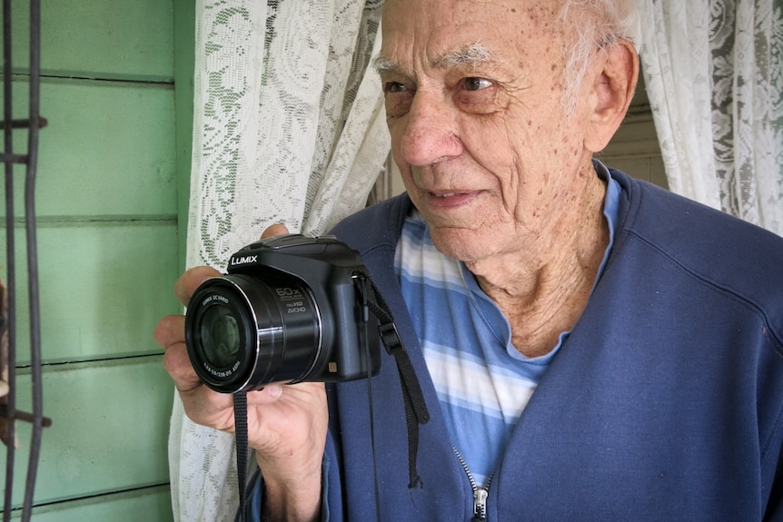 An elderly man holds a camera in one hand, a window with lace curtains behind him.