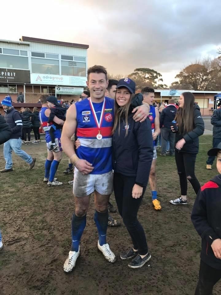 A man in muddy footy gear smiles while hugging his female partner, dressed in club merchandise. 