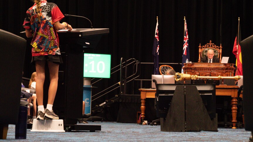 Kid wearing school uniform on plastic stool inside a convention centre at a mock-up parliament sitting.