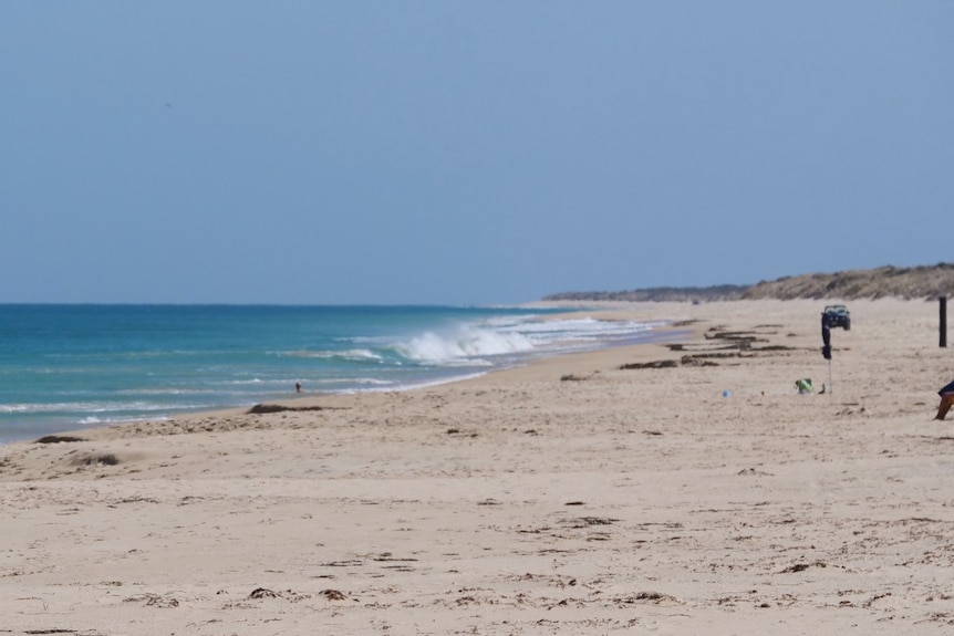 Stretch of beach with white sand and appealing blue water, with people walking on it in the distance.