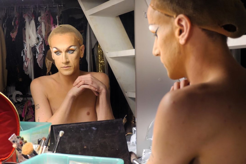 A drag queen, not yet in costume, in front of a mirror backstage.