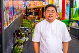 A man in a white chef jacket stands smiling in a colourfully painted lane.