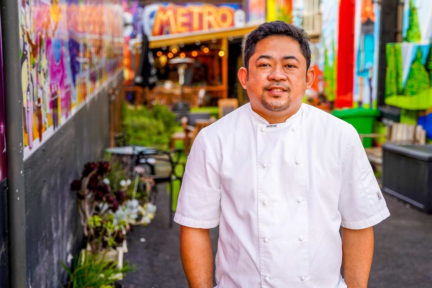 Un homme vêtu d'une veste de chef blanche se tient en souriant dans une ruelle peinte de couleurs vives.