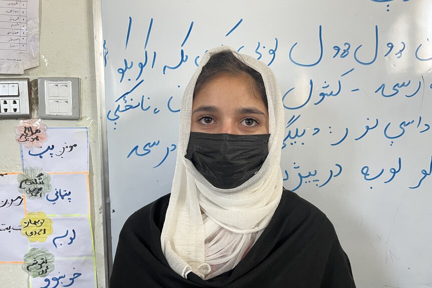 A girl in white hijab and black face mask with Arabic writing on the whiteboard behind her. 
