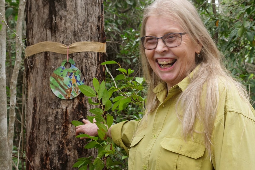 Margaret in a green shirt and glasses leans on the tree where her art is hanging.
