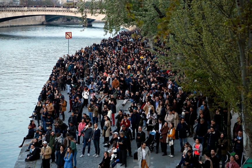 A large crown of people gather along a riverbank to watch a fire burn through a cathedral