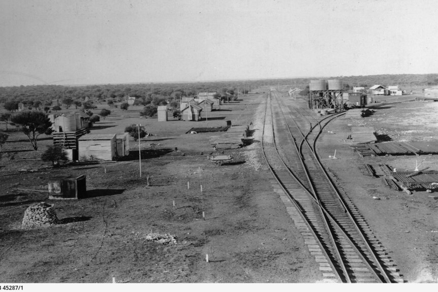 Black and white photograph showing desert landscape with railway tracks and smattering of huts alongside tracks