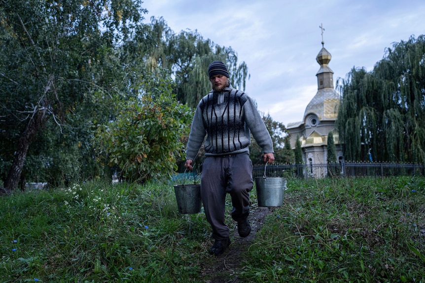 A young man in grey, winter civilian clothing carries two silver buckets in front of a church.