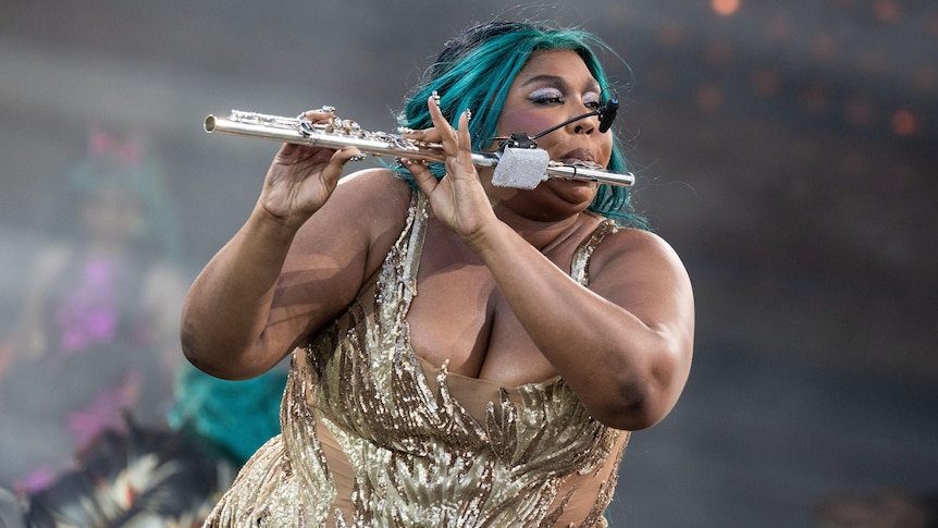 Lizzo plays a silver flute on stage. There is a microphone clipped to the flute, she has sea-green hair and wears gold.