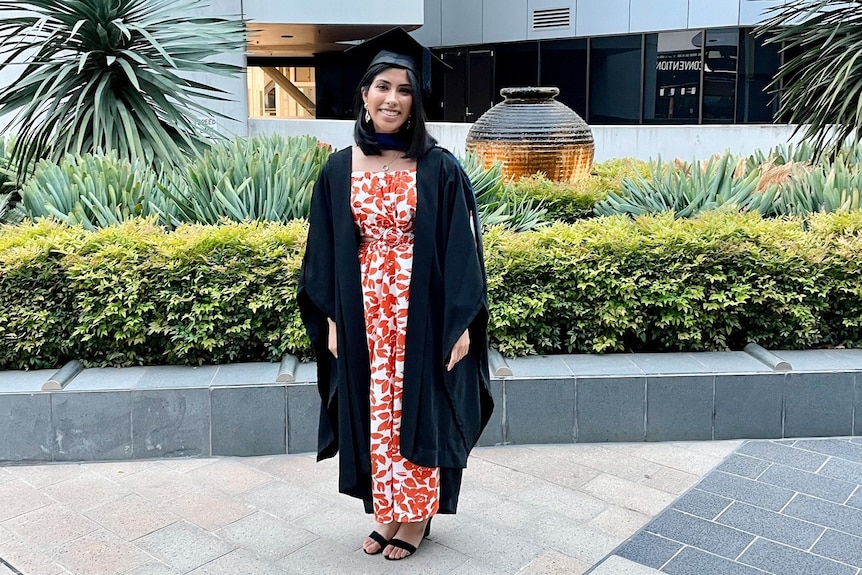 A woman in a graduation gown and hat stands in front of a large university building smiling.