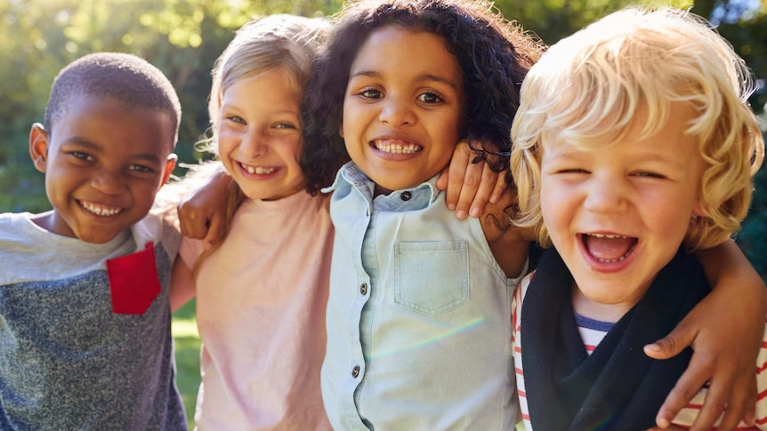 Four children of diverse backgrounds smile with their arms draped around one another in a sunny garden.
