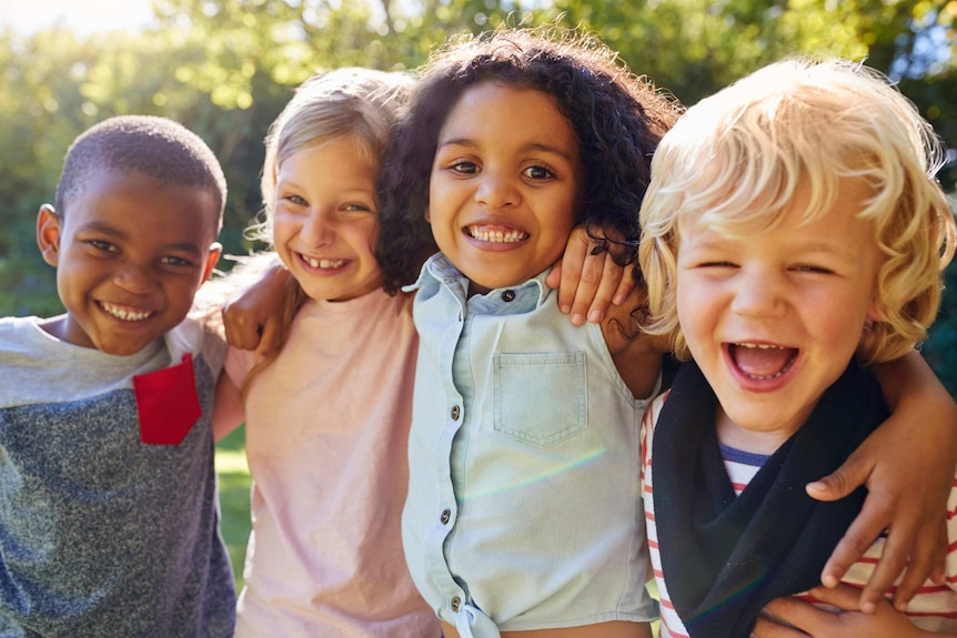 Four children of diverse backgrounds smile with their arms draped around one another in a sunny garden.