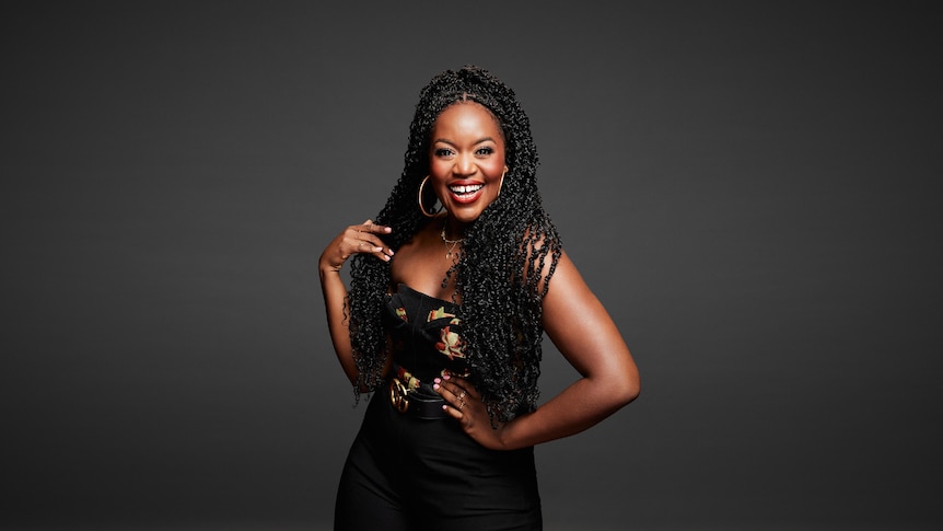 A broadly smiling woman with braids and large hope earrings in a dark jumpsuit