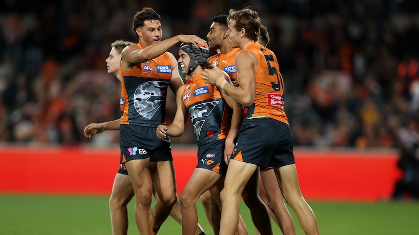 GWS players walk back after a goal, patting a smiling teammate who is wearing orange and black headgear.