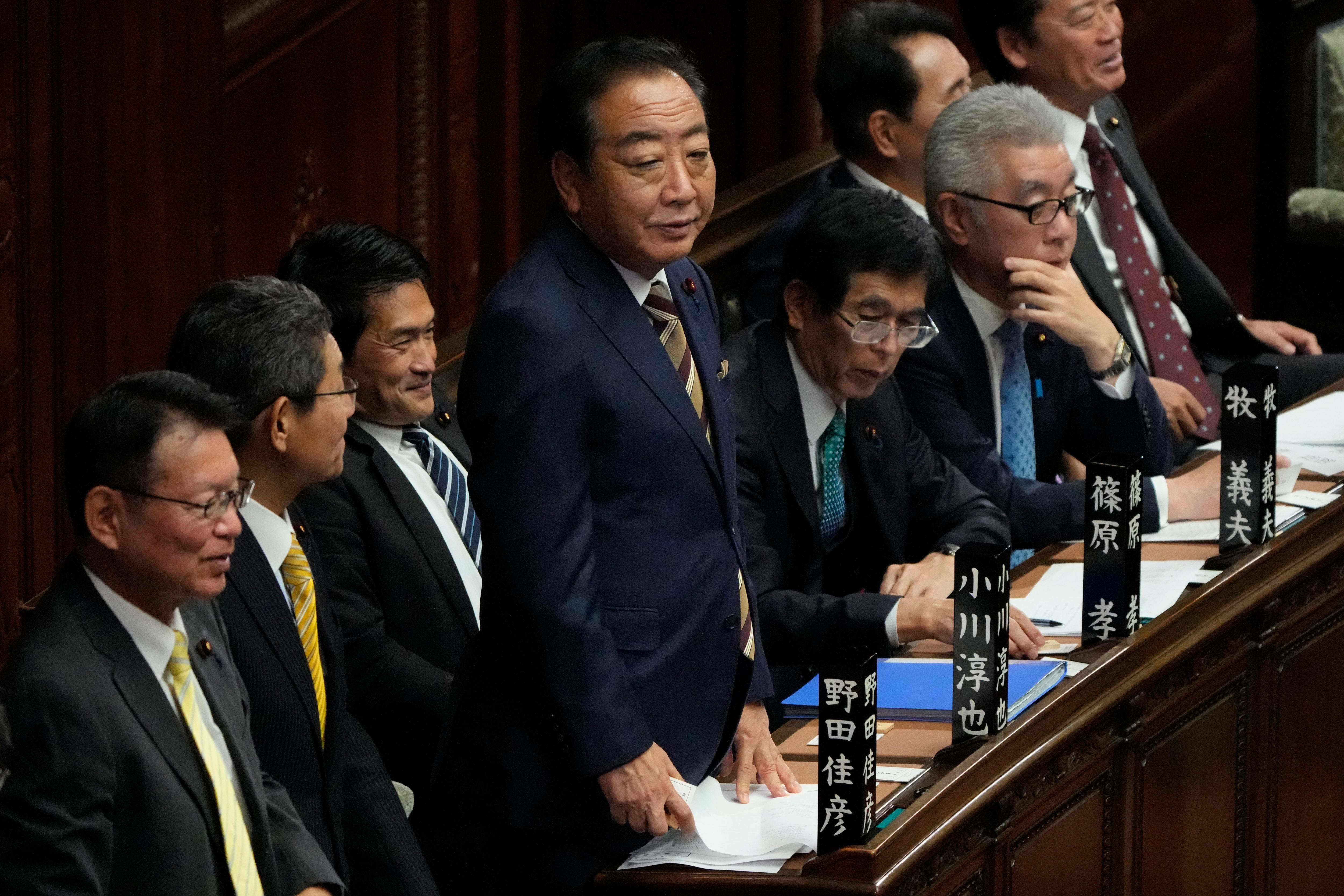 Yoshihiko Noda stands in Japanese parliament. He is next to a number of other seated lawmakers.