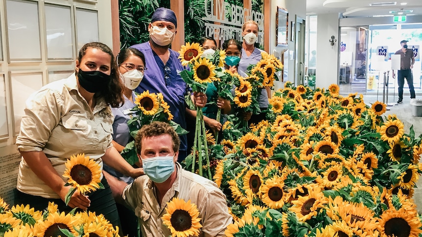 Two farmers and hospital staff pose with many sunflowers in a hospital