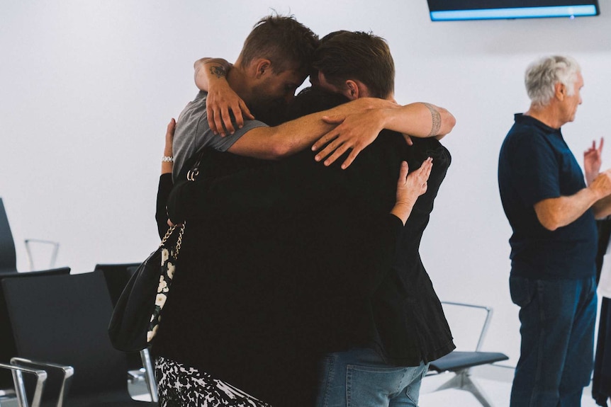 Three people hug at an airport departure lounge as two others talk in the background.