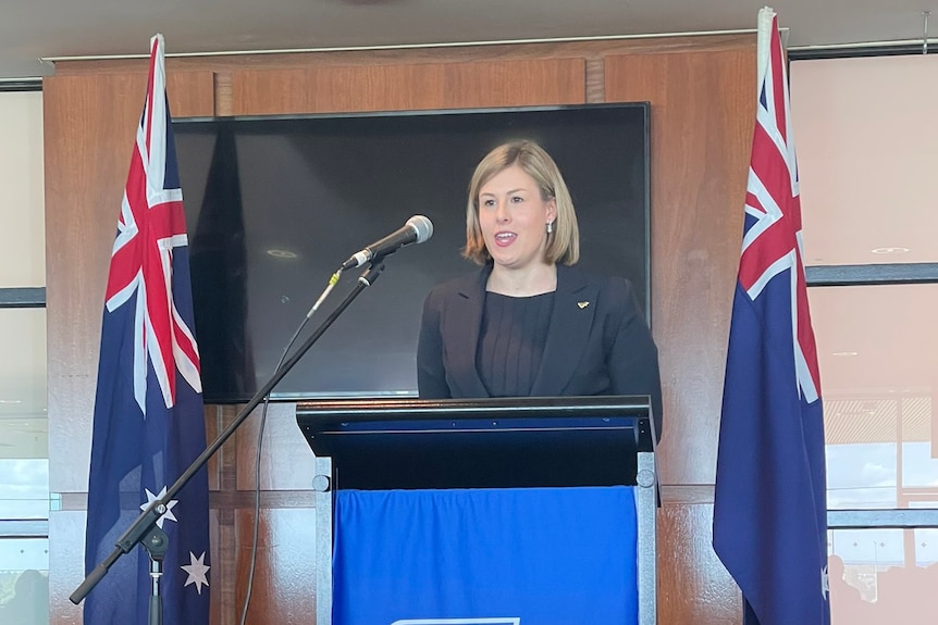 Une femme portant un blazer debout sur un podium flanqué de deux drapeaux australiens.