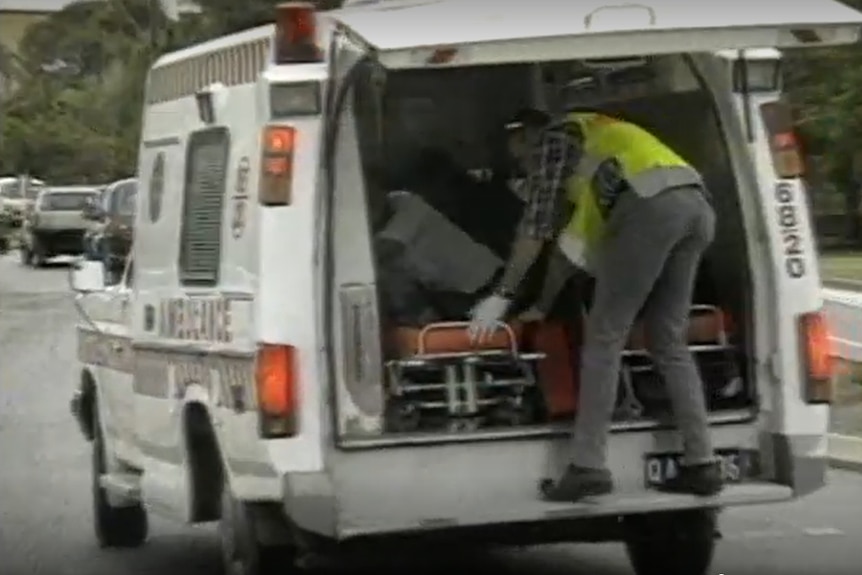An old ambulance being driven with its rear door up, with a person attending to an injured bikie.