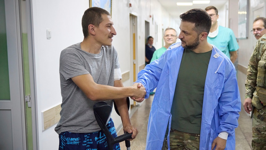 ukrainian president volodymyr zelensky wearing surgical gown over clothes shakes hands with soldier on crutches missing one leg