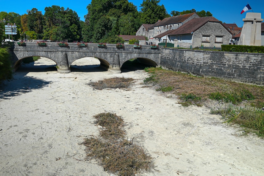 A dried-up river bed with no water passes under a low three arch bridge.