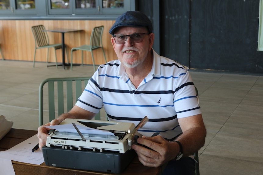 An older man in a striped polo shirt smiles while holding a retro typewriter.