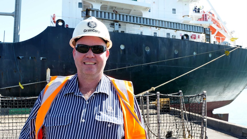 Jeff Moodie smiles at the camera wearing a high-visibility vest and hard helmet, standing in front of a large ship.