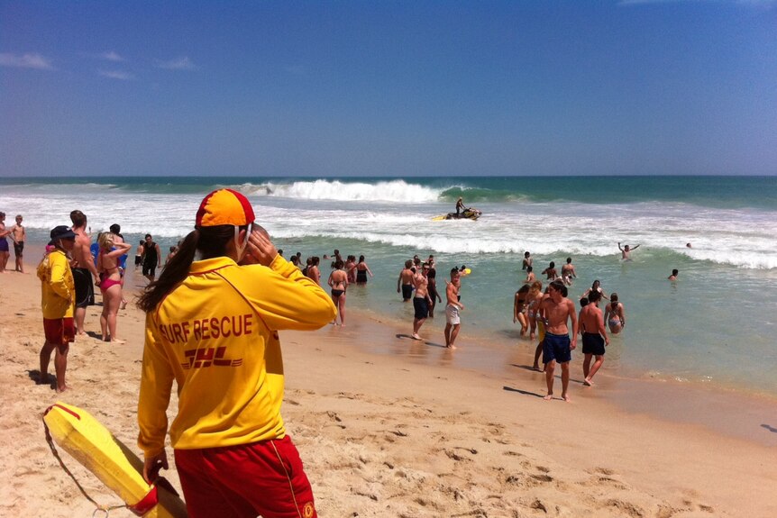 A surf life saver looks out at the ocean on a sunny day surrounded by beachgoers.