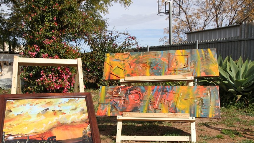 A framed oil painting and two colourful abstract works sit on easels outside