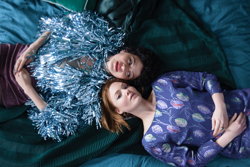 Two women, one wearing a bright coloured tassel jacket the other a patterned dress, lie on their backs on bed with heads close.