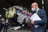 Masked man with file in hand standing in front of reconstructed airplane wreckage.