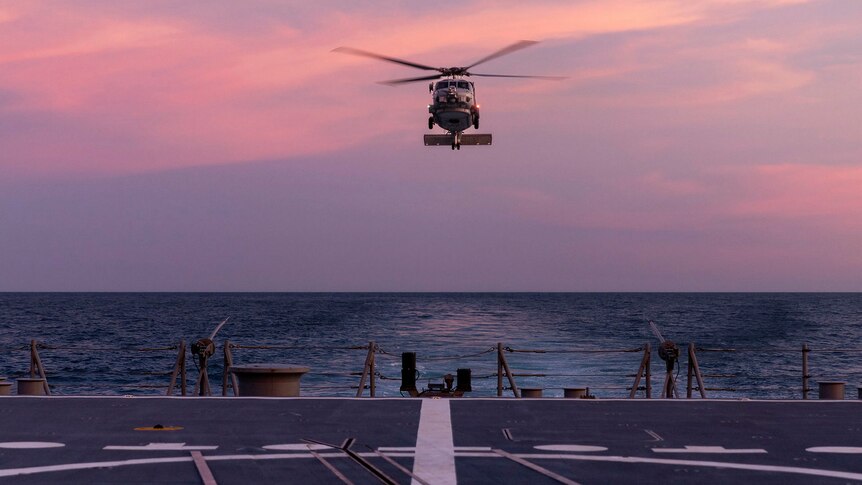 A helicopter lands on a warship's deck at sunset.