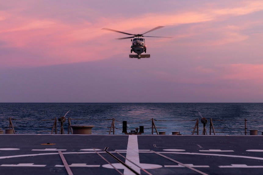 A helicopter lands on a warship's deck at sunset.