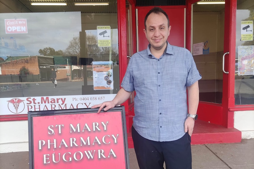 A smiling middle-aged man wears blue shirt, pants, standing next to a red sign saying St Mary Pharmacy Eugowra.