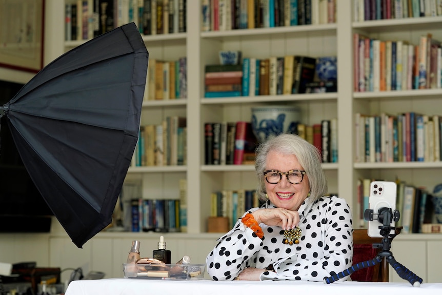 Older woman smiling with a phone on tripod and miniature studio lightening equipment in front of her