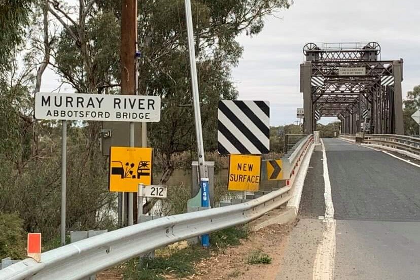 A sign saying "Murray River Abbotsford Bridge" next to traffic signs and a road leading onto a metal bridge.