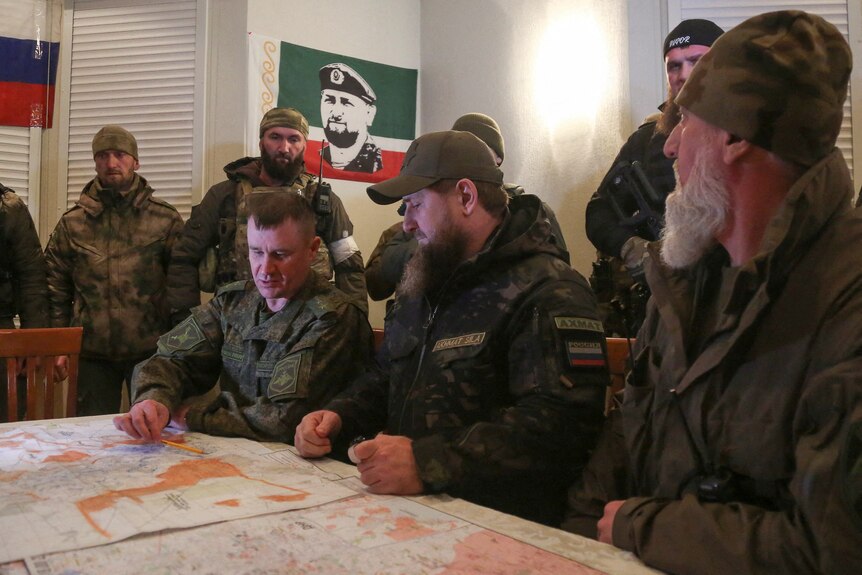 A group of men in camo gear sit around a table looking and pointing at maps