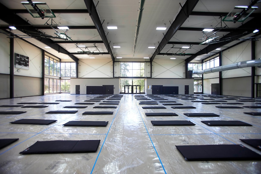 Black mats laid in rows on a gymnasium floor. Plastic wrap and tape marks sections around each mat. AC on ceilings