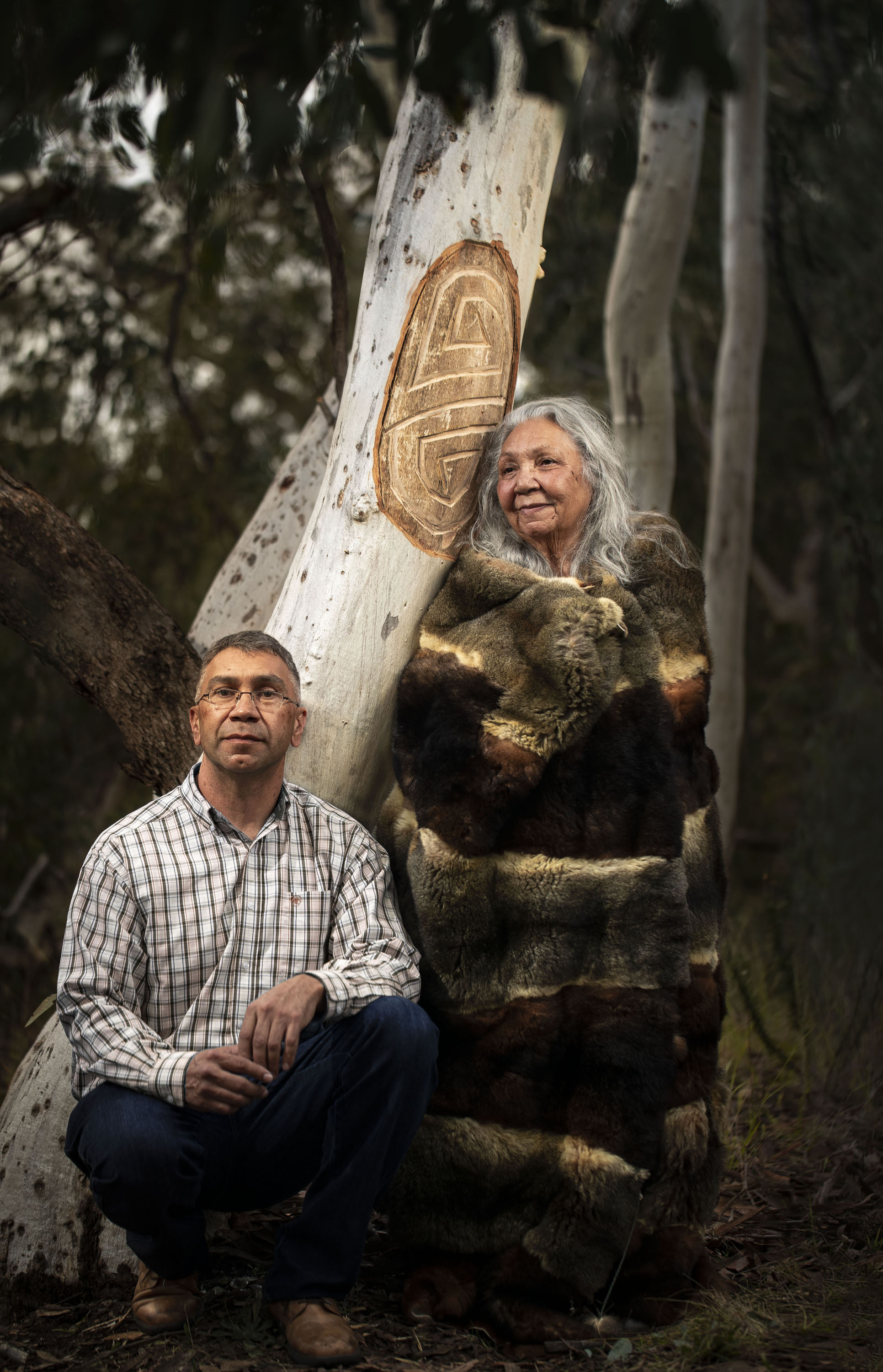 A grizzled aboriginal man squats next to an older aboriginal woman standing next to a tree that has been carved with a design
