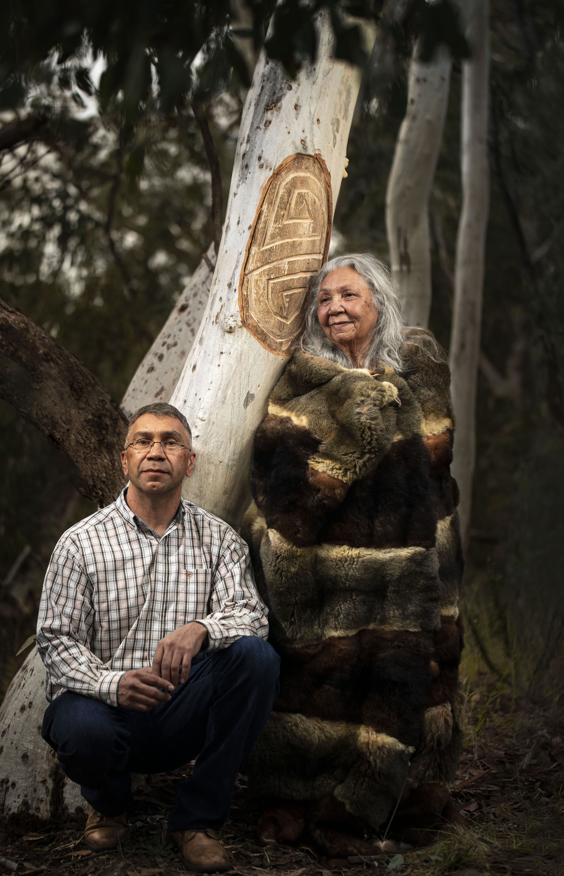 A greying Aboriginal man crouches beside an older Aboriginal woman standing beside a tree that has been carved with a design