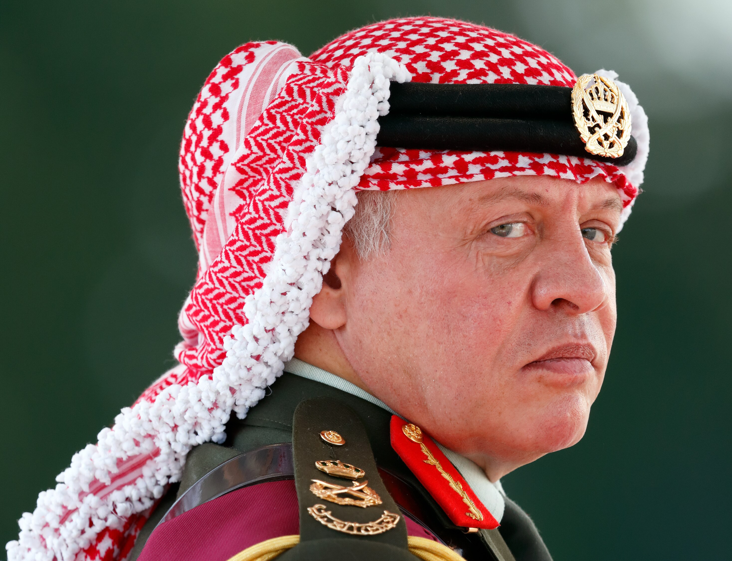 King Abdullah II looks over his shoulder at the camera in a close-up shot. His appearance is stern he wears military attire.